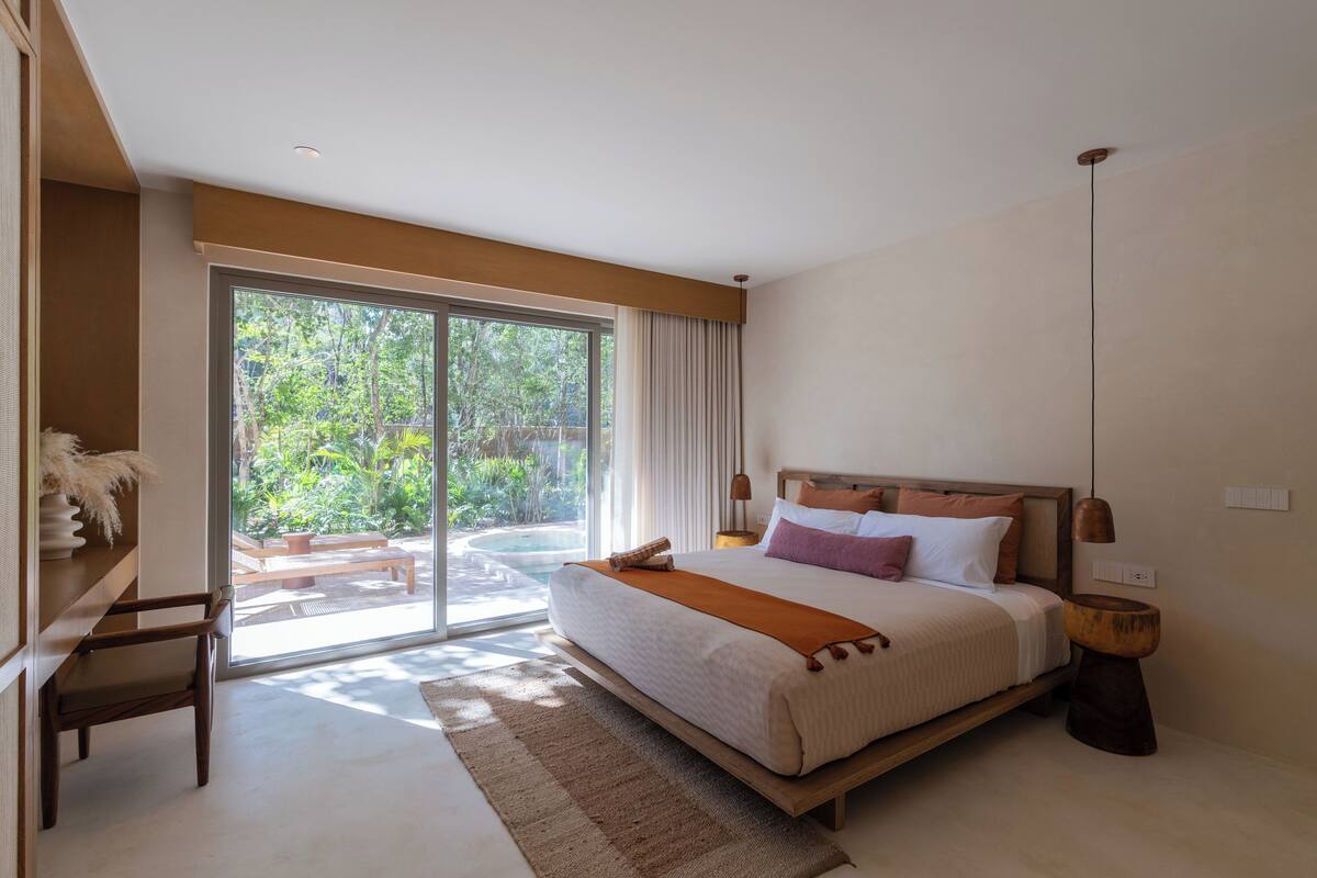 Quarto com uma cama grande, mesa de cabeceira de madeira, uma cadeira e vista para uma piscina e vegetação lá fora.
