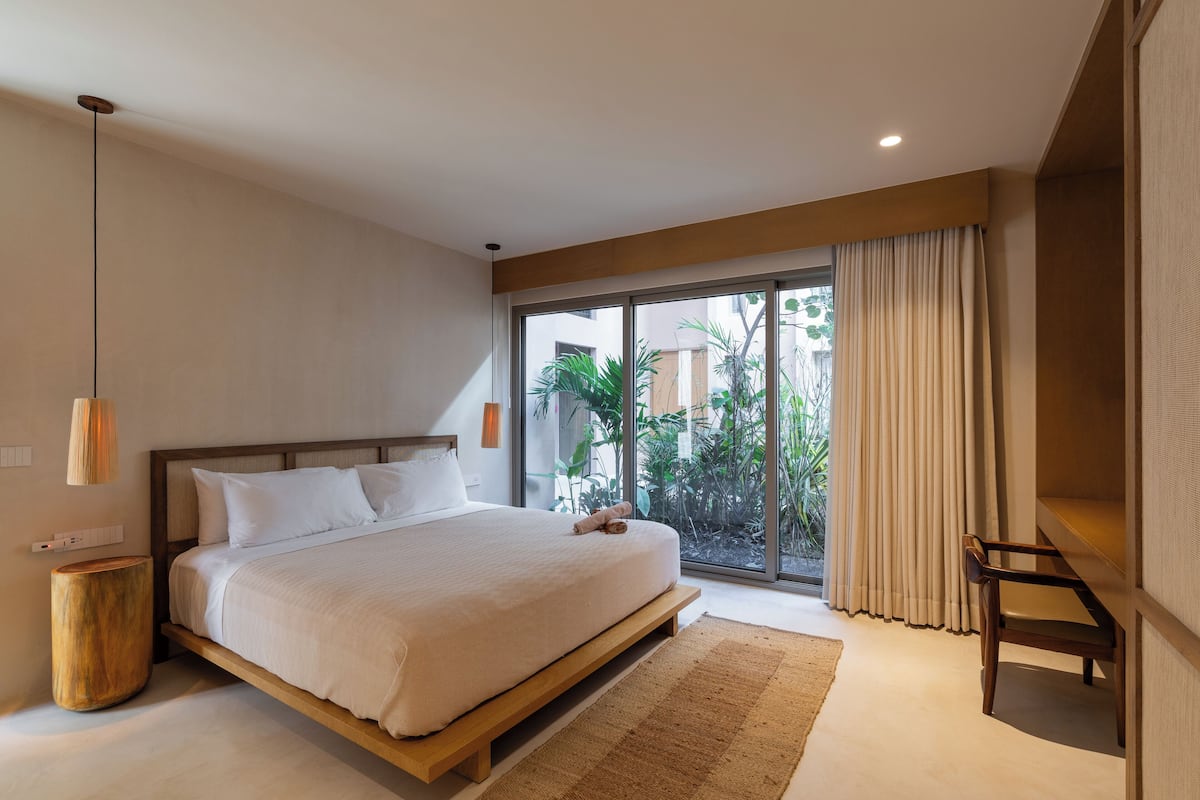 Quarto moderno com cama grande, mesa de cabeceira de madeira, luminária pendente e vista para o verde pela janela.