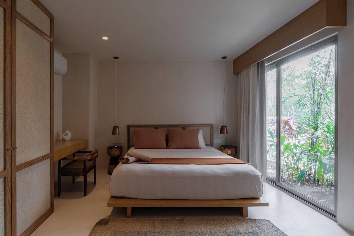Quarto moderno com uma cama grande, uma mesinha de cabeceira de madeira, uma pequena mesa e vista para o verde pela janela.
