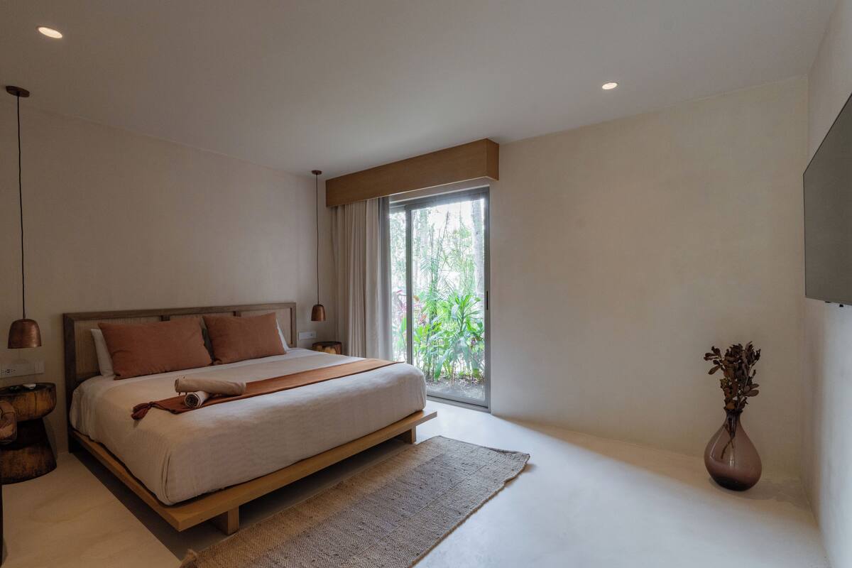 Quarto moderno com uma cama grande, mesa de cabeceira, abajur e janela com vista para o verde.