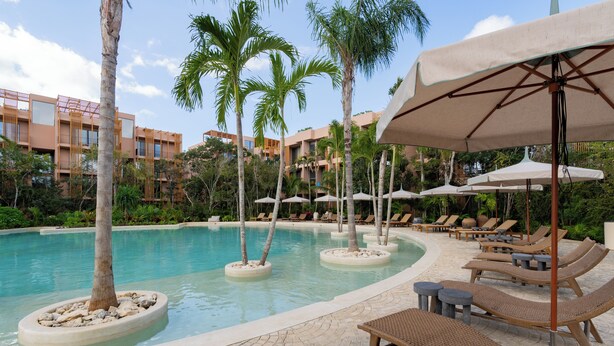 A pool area with lounge chairs and umbrellas, surrounded by palm trees and a building with balconies. - Aflora Tulum  (Tulum)