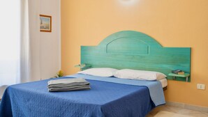Ein Bett mit blauer Bettdecke und einem türkisblauen Kopfteil, ein kleiner Nachttisch mit einem Buch sowie eine Wandleuchte.