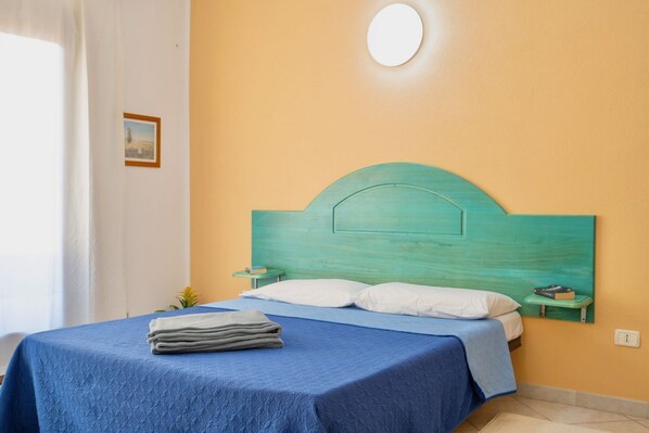 A bed with a blue bedspread and a teal headboard, a small bedside table with a book, and a wall-mounted light fixture.