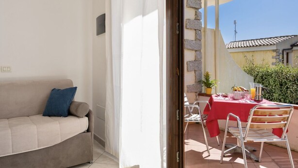 Ein Balkon mit einem roten Esstisch, einer weißen Couch und Blick auf ein Gebäude mit einem Ziegeldach.