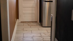 A narrow kitchen with a white door, a stainless steel refrigerator, and a window with a view of a wooden structure outside.