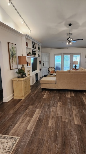 A living room with a wooden floor, a sofa, a television, and a ceiling fan. - Designer's Gorgeous Lake House with King Bed on Lake Tarpon (Palm Harbor)