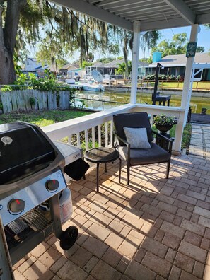 A covered patio with a grill, a chair, and a view of a canal with boats. - Designer's Gorgeous Lake House with King Bed on Lake Tarpon (Palm Harbor)