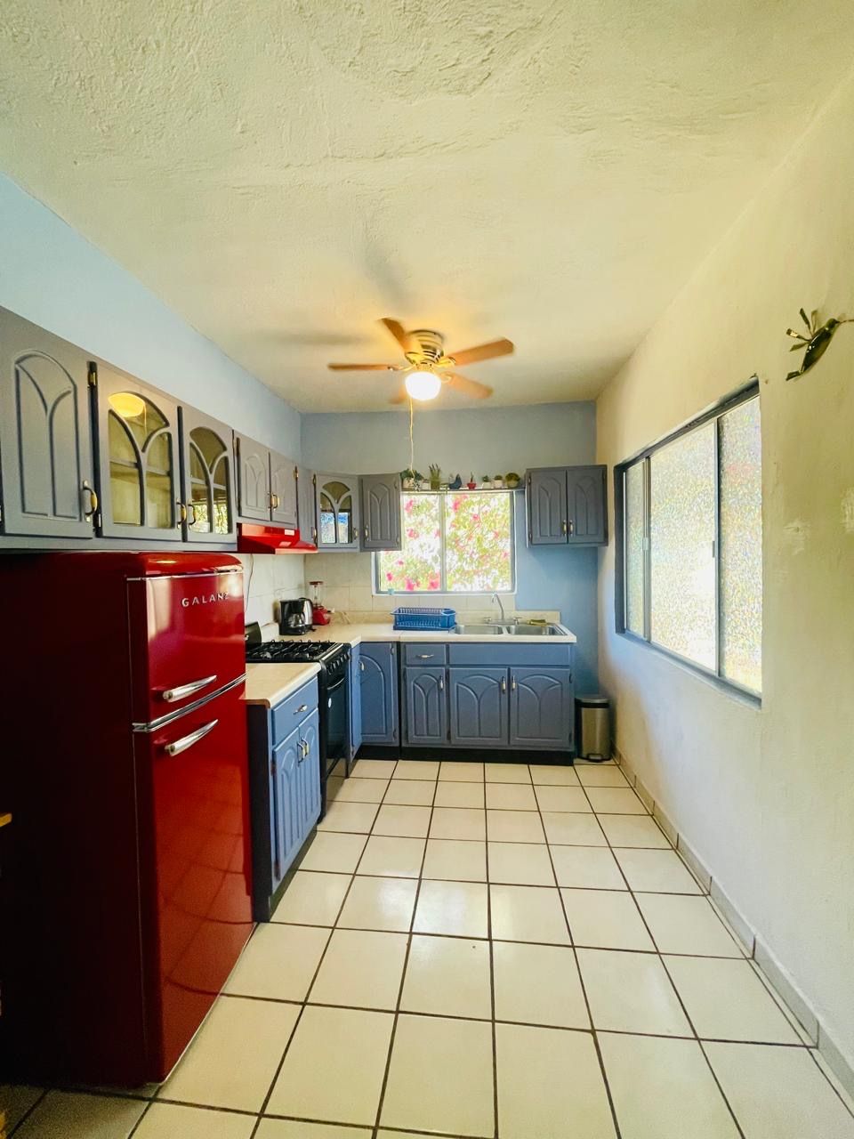 A compact kitchen with a red refrigerator, blue cabinets, a ceiling fan, and a window with a view.