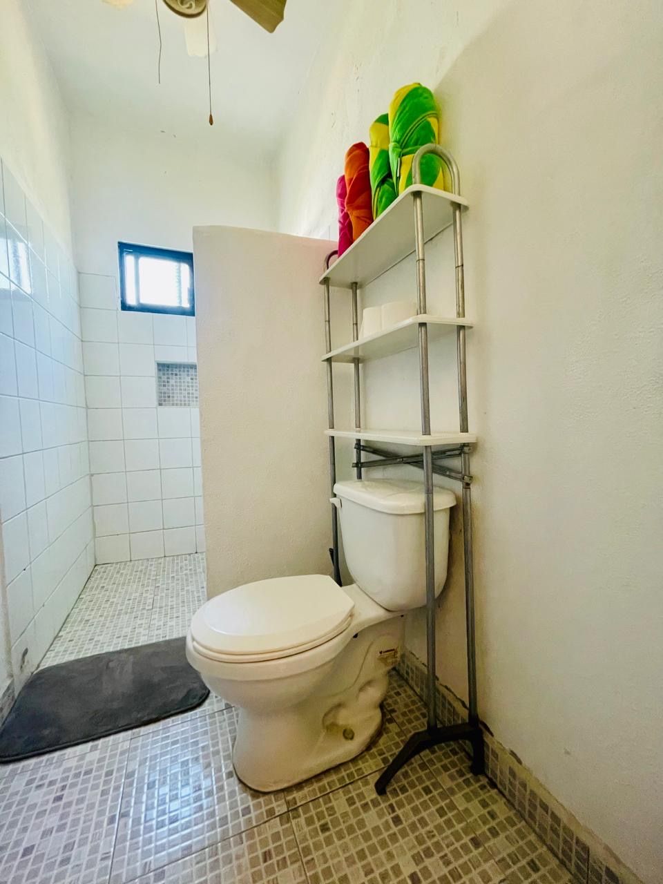 A small bathroom with a toilet, a shelf with colorful items, and a window.