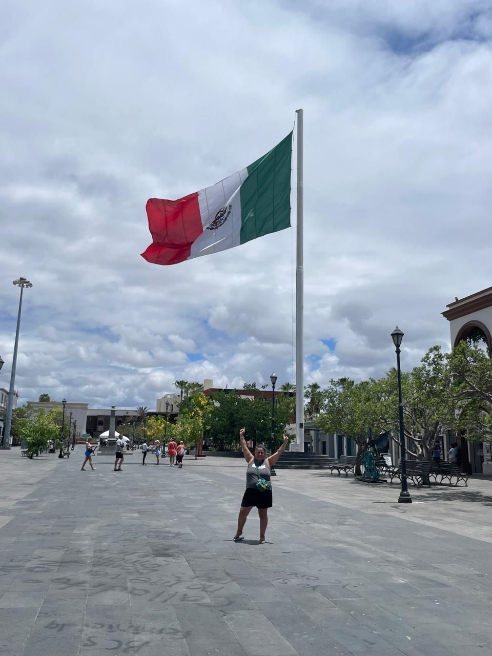 Un asta con una bandera, una plaza empedrada y gente caminando.