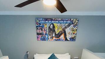 A room with a bed, a ceiling fan, a wall with a colorful poster, and a small table.