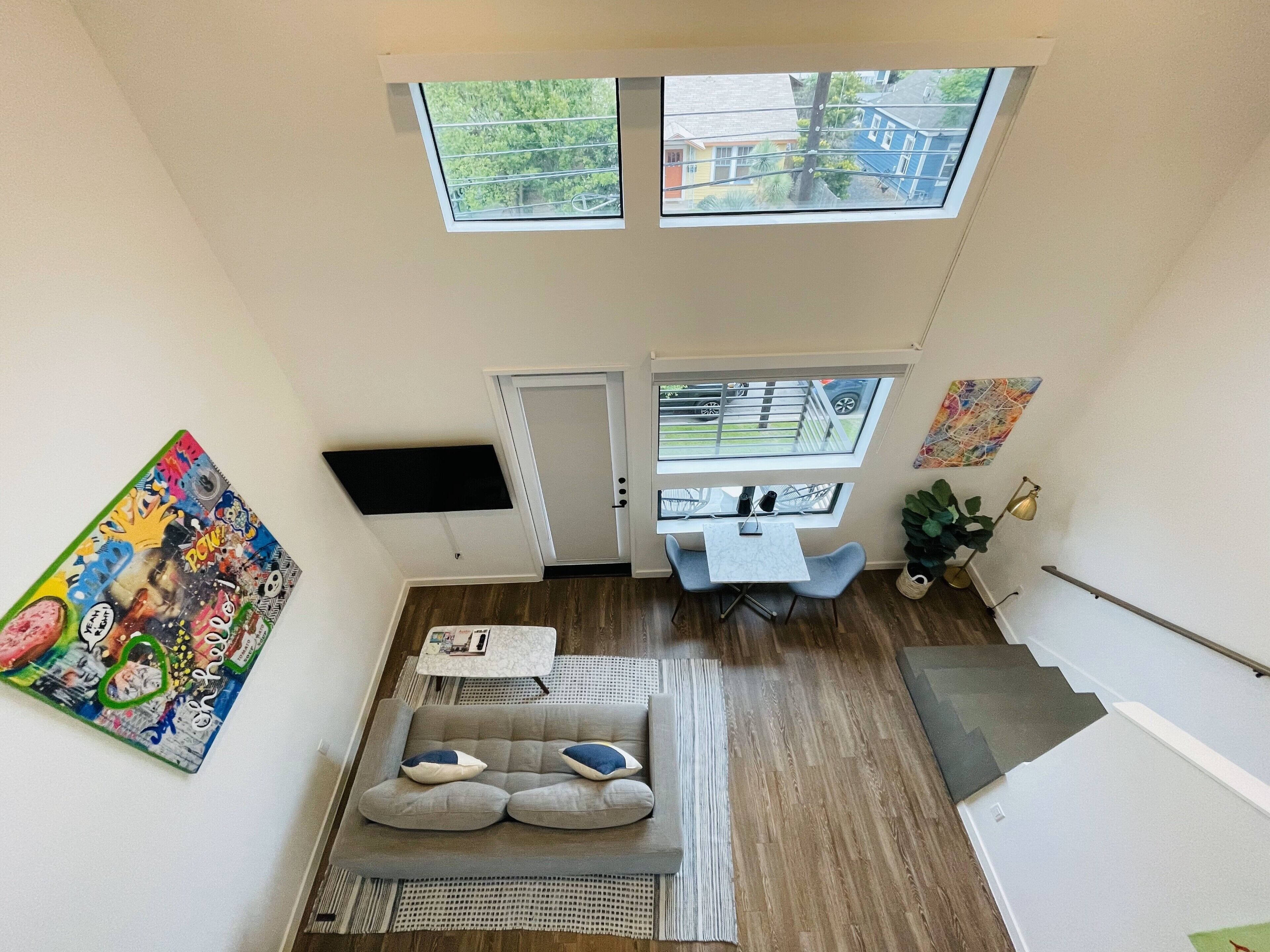 A compact living space with a sofa, a small table, and a TV mounted on the wall. There are two windows providing natural light and a view of the outside.