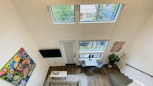 A compact living space with a sofa, a small table, and a TV mounted on the wall. There are two windows providing natural light and a view of the outside. - NEW Cozy Downtown East Austin Condo - Prime Location! 10 min walk to Rainey St (Austin)