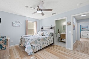 Une chambre à coucher comprenant un lit, un ventilateur de plafond, une fenêtre avec des rideaux, une table de chevet et une commode.