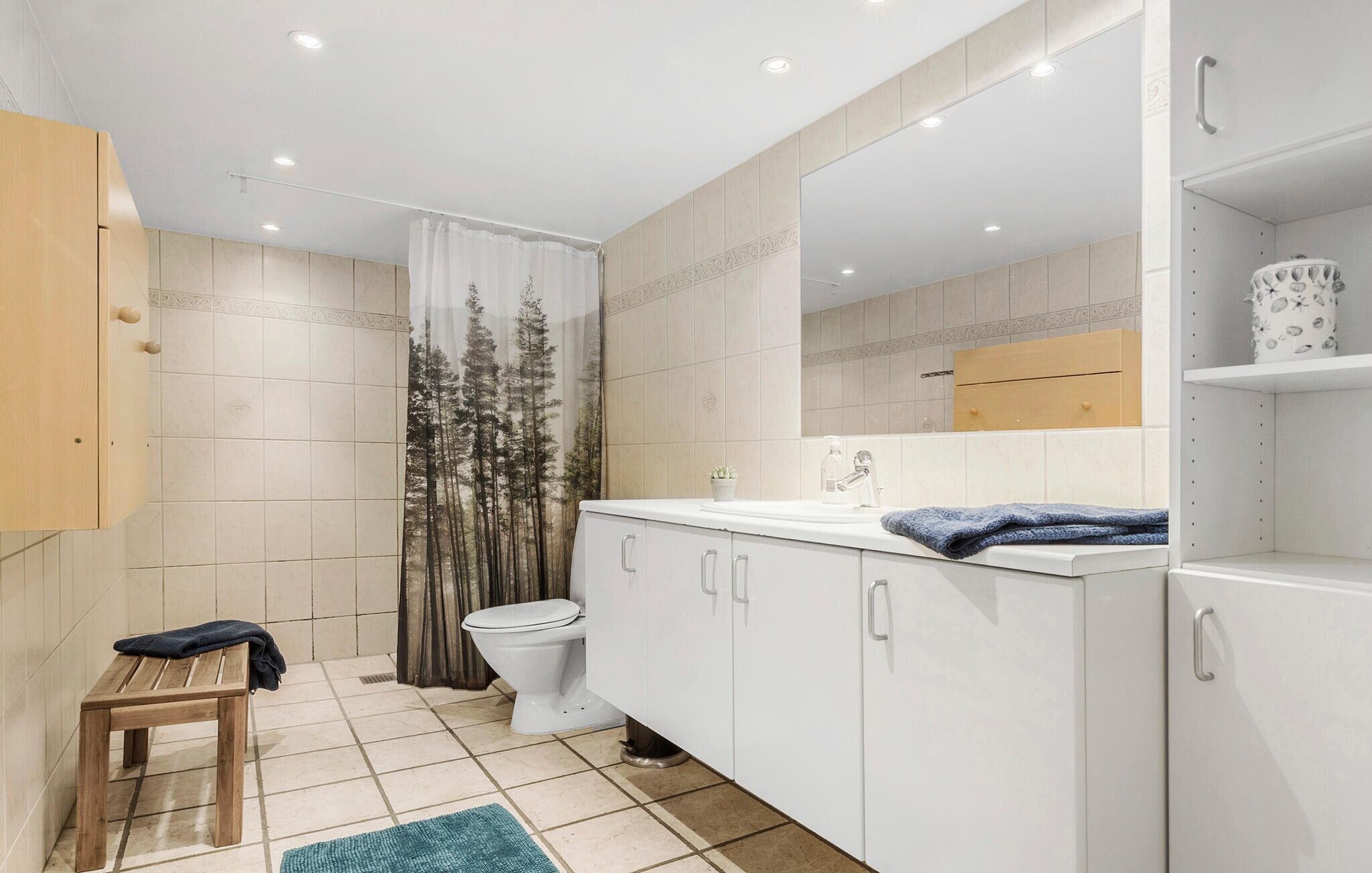 Moderni kylpyhuone, jossa on wc, pesuallas ja peili. Pesualtaan yläpuolella on metsäkuviollinen suihkuverho, pieni jakkara ja sininen pyyhe.