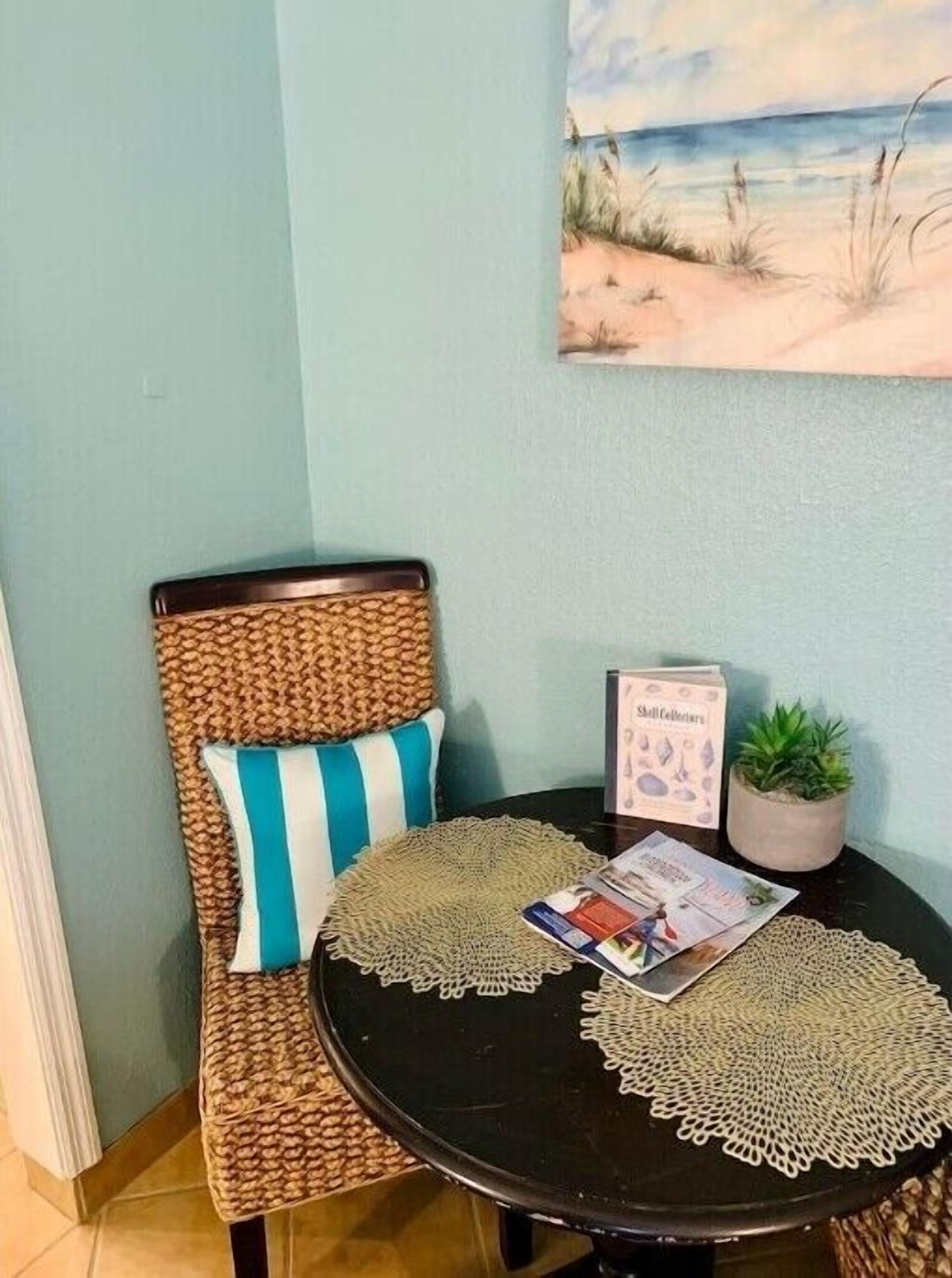 Un rincón de una habitación con una silla de mimbre, una mesita redonda con un mantel individual, una planta en maceta y un cuadro en la pared.