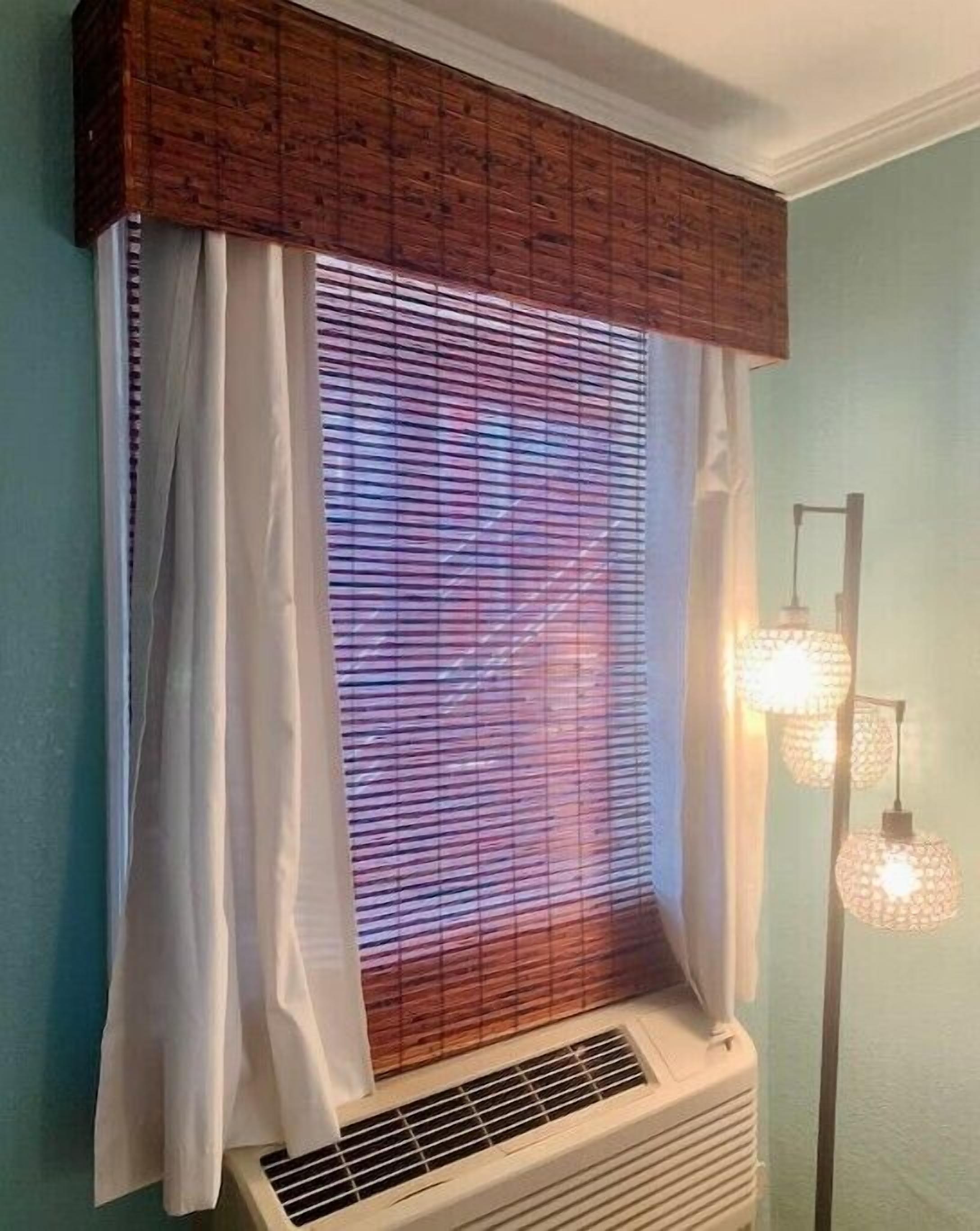Une pièce avec une fenêtre recouverte de stores et de rideaux légers, une grille d’aération située sous la fenêtre et un luminaire fixé au mur.