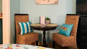 Une pièce comprenant deux fauteuils en osier, une petite table ronde et un tableau représentant une scène de plage accroché au mur.