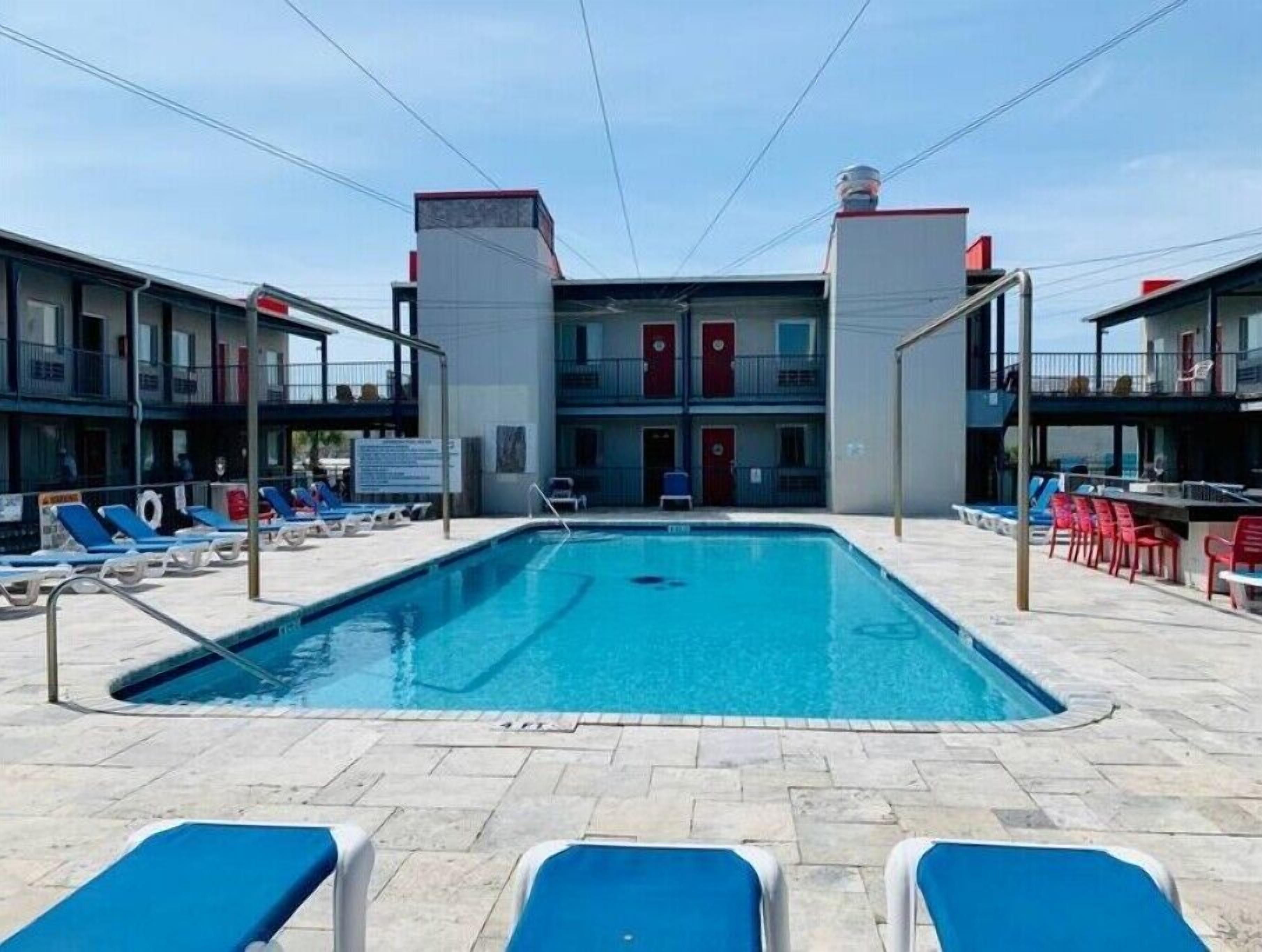 A swimming pool with blue lounge chairs, a building with red doors, and a clear sky.