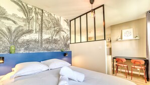 A neatly made bed with white linens and rolled towels, a blue headboard, a wall mural of tropical foliage, a desk with two orange chairs, and a hanging light fixture. - Le Verde - Ravissant appartement cosy (Saint-Genis-Laval)