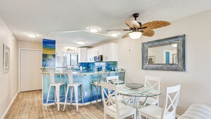 Une cuisine avec un coin bar, des tabourets de bar blancs, une table à manger avec des chaises et un ventilateur de plafond.