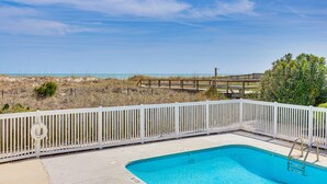 Une piscine entourée d’une clôture blanche, avec une bouée de sauvetage et une terrasse en bois qui mène à la plage.