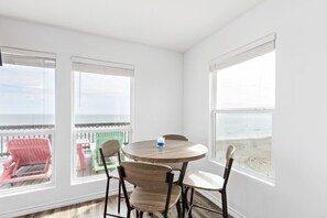 A room with a round wooden table and four chairs, large windows with white blinds, and a view of the beach.
