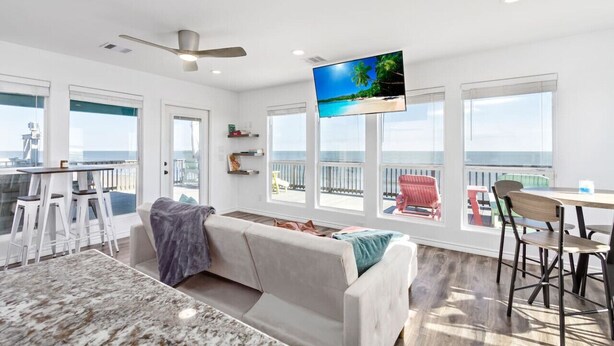 A spacious living area with a large TV, a balcony with a view of the ocean, and a dining area with a table and chairs. - Cabn By the Sea Ultra Secluded Beachfront Home (Bolivar Peninsula)