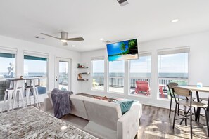 A spacious living area with a large TV, a balcony with a view of the ocean, and a dining area with a table and chairs.