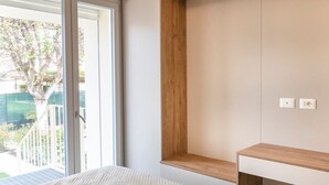 Une chambre avec une grande fenêtre, une étagère en bois et une table de chevet blanche.