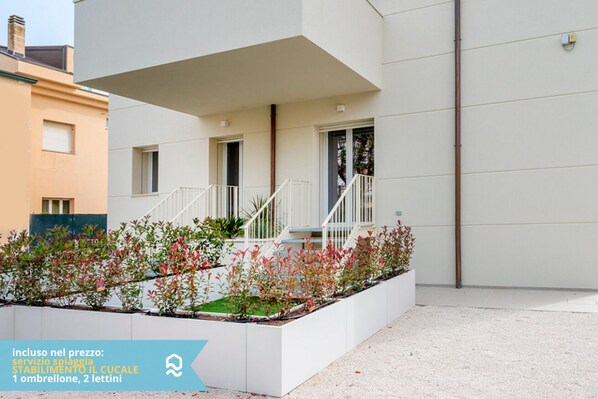 A modern building with a white facade, a balcony with white railings, and a garden area with various plants.