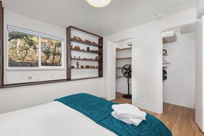 Une chambre avec une grande fenêtre, un lit recouvert d’une couverture turquoise et une étagère intégrée où sont disposés des objets décoratifs.