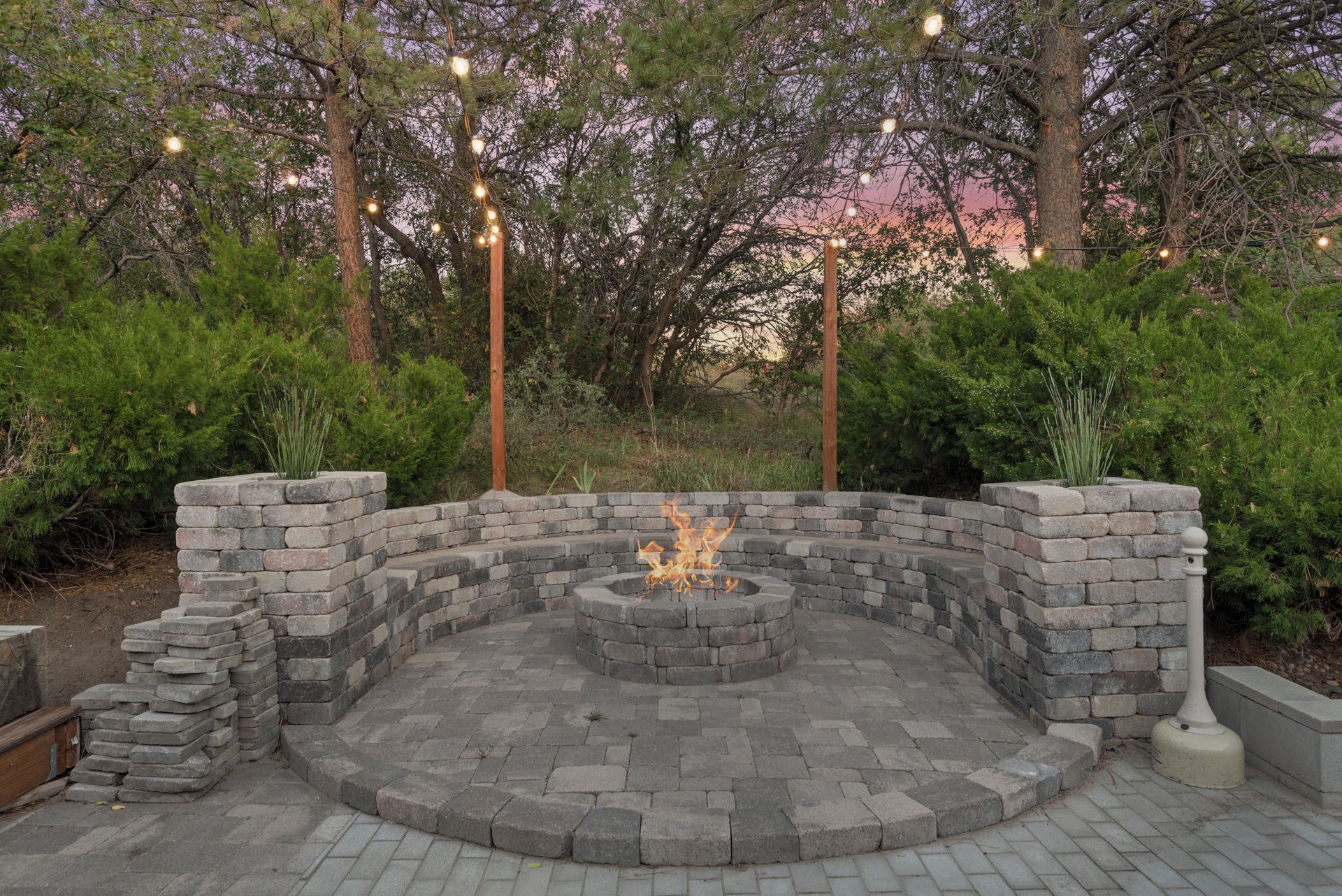 A circular stone fire pit surrounded by a low stone wall, with string lights above and dense shrubbery in the background.