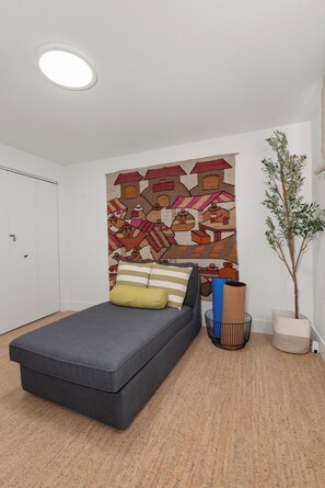Un dormitorio moderno con un sofá cama gris, un tapiz de colores, una planta en maceta y una puerta blanca.