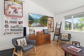 A living room with a television, a wooden cabinet, a chair with a cushion, a small round table with books, and a window with a view of trees. - Wilderness Haven with Hot Tub in Franktown (Franktown)