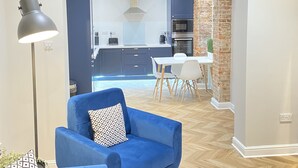 A modern living room with a blue armchair, a dining area with a white table and chairs, and a kitchen with dark cabinets and a brick accent wall. - Waimea Apartment 1| Best Location, Beach Front, Parking, Scarborough (North Yorkshire)