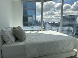 Ein Schlafzimmer mit großem Fenster, einem Bett mit weißer Bettwäsche und Blick auf eine Stadtlandschaft.