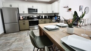 Una cuina moderna amb armaris blancs, electrodomèstics d'acer inoxidable i una zona de menjador amb taula per a dues persones.