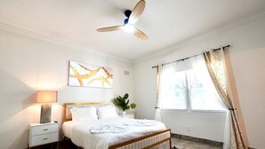 A bedroom with a bed, bedside tables, a ceiling fan, a window with curtains, and a wall-mounted artwork. - SōLE Villa #2|Elegant 2BR Retreat|Beach|Stargazing (Cocoa Beach)
