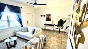 A living room with a white sofa, a small round table, a striped armchair, a ceiling fan, a large plant, and a painting on the wall. - SōLE Villa #2|Elegant 2BR Retreat|Beach|Stargazing (Cocoa Beach)