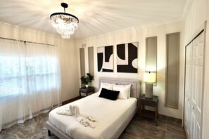 A hotel room with a large bed, a chandelier, a nightstand with a lamp, and abstract wall art. - Villa Fiorō-Bold Luxury 2BR /River & Coastal Vibe (Cocoa Beach)