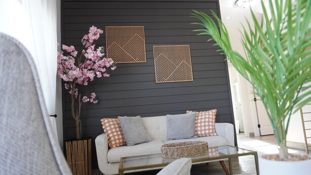 Un salon moderne avec un mur en bois foncé, un canapé blanc agrémenté de coussins à motifs, une table basse en verre et des panneaux décoratifs en bois.
