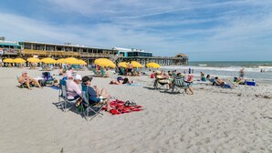 Une plage où des gens se détendent sous des parasols, une jetée et un bâtiment avec un toit bleu.