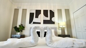 Une chambre d’hôtel avec un lit, deux serviettes disposées en forme de cœur, une plante en pot et des œuvres d’art abstraites accrochées au mur.