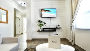 A modern living room with a flat-screen TV mounted on the wall, a round coffee table, a sofa, a potted plant, and a small shelf with decorative items. - Luxury 2BD 2BTH - Condo #5 (Cocoa Beach)