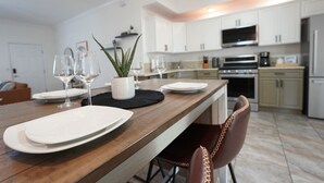 A modern kitchen with white cabinets, a wooden dining table, and a black placemat. - Luxury 2BD 2BTH - Condo #5 (Cocoa Beach)