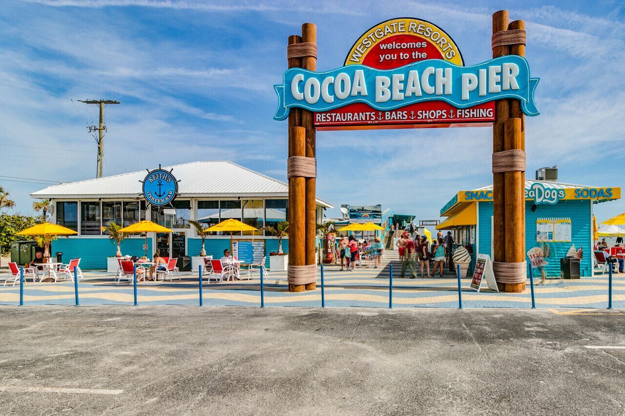 A beach pier with a sign reading "Cocoa Beach Pier" and "Westgate Resorts Welcome to the Cocoa Beach Pier Restaurants, Bars, Shops, and Fishing." There are outdoor seating areas with yellow umbrellas, and a building with a blue and white facade in the background.