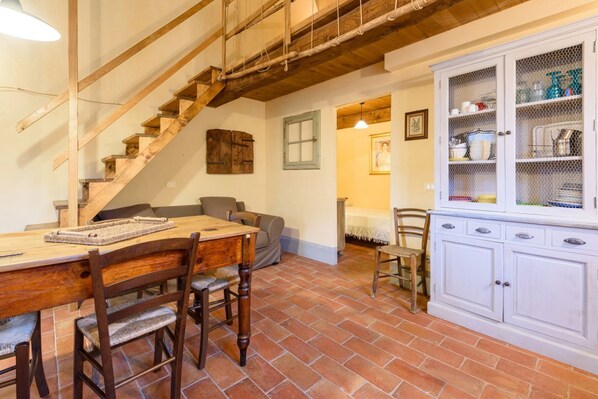 A rustic interior with a wooden staircase, a dining area with a wooden table and chairs, a white cabinet with glass doors, and a tiled floor.