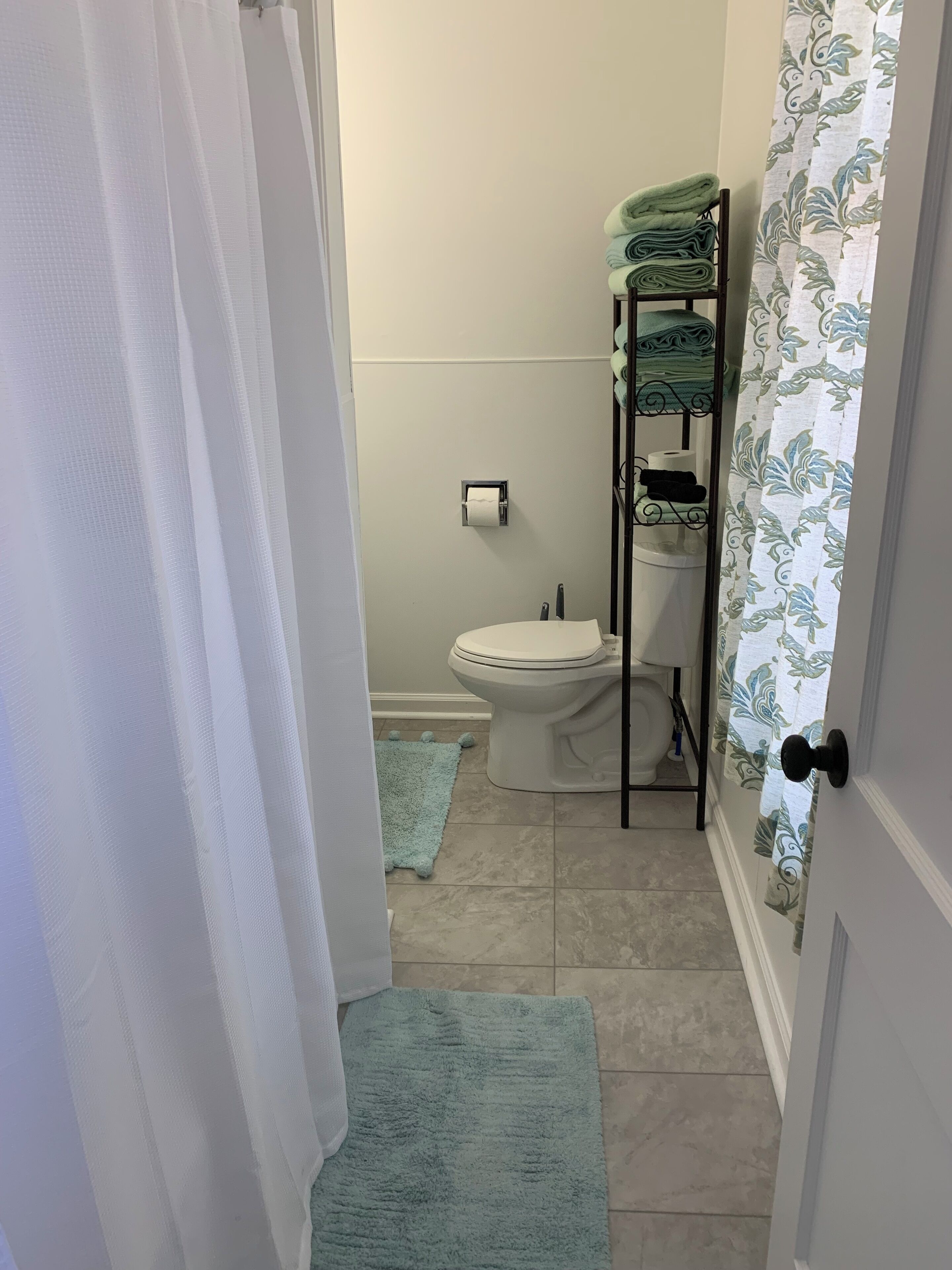 A bathroom with a toilet, a towel rack with folded towels, a toilet paper holder, and a small blue rug on the floor.