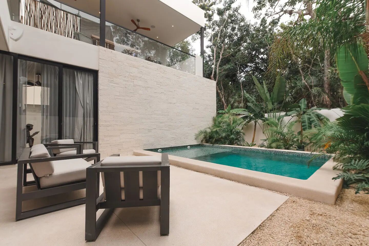 Zona de piscina exterior moderna amb un disseny minimalista, que inclou una zona de seients amb una cadira i una taula, envoltada de vegetació exuberant i una piscina nítida.
